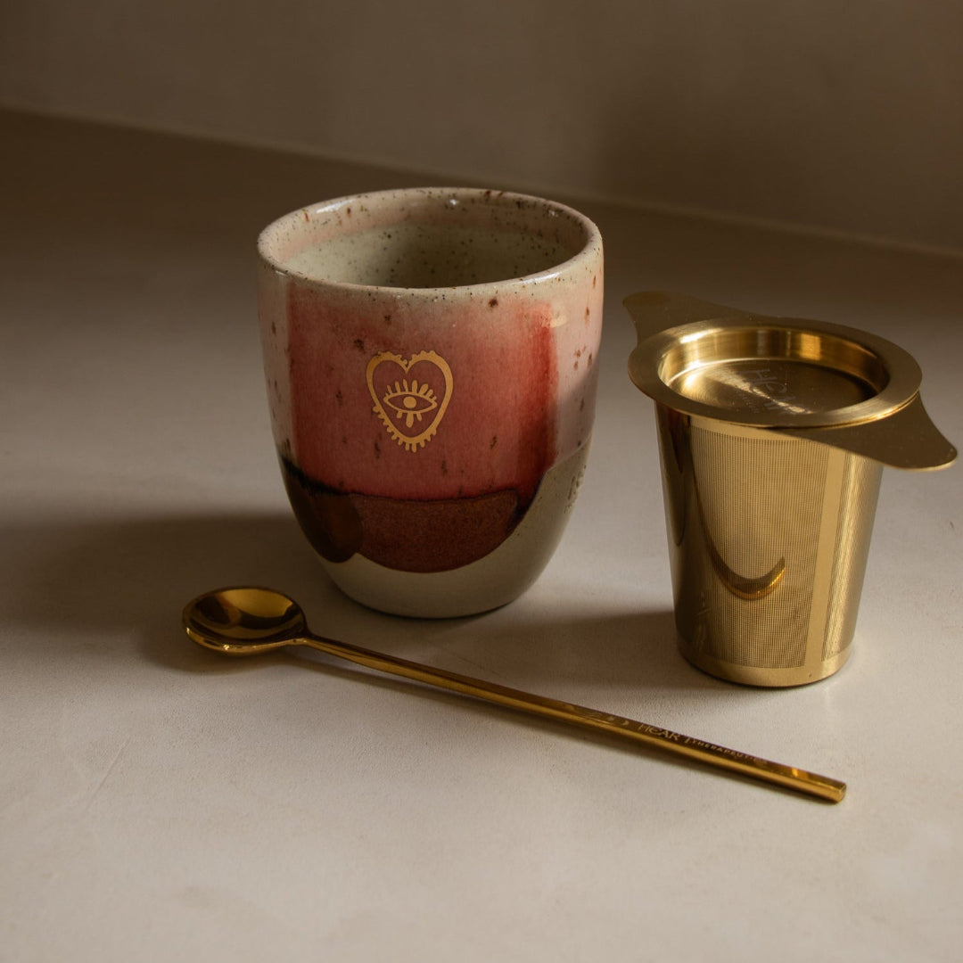 Ceramic mug with gold heart design, gold spoon, and gold cup on a neutral background