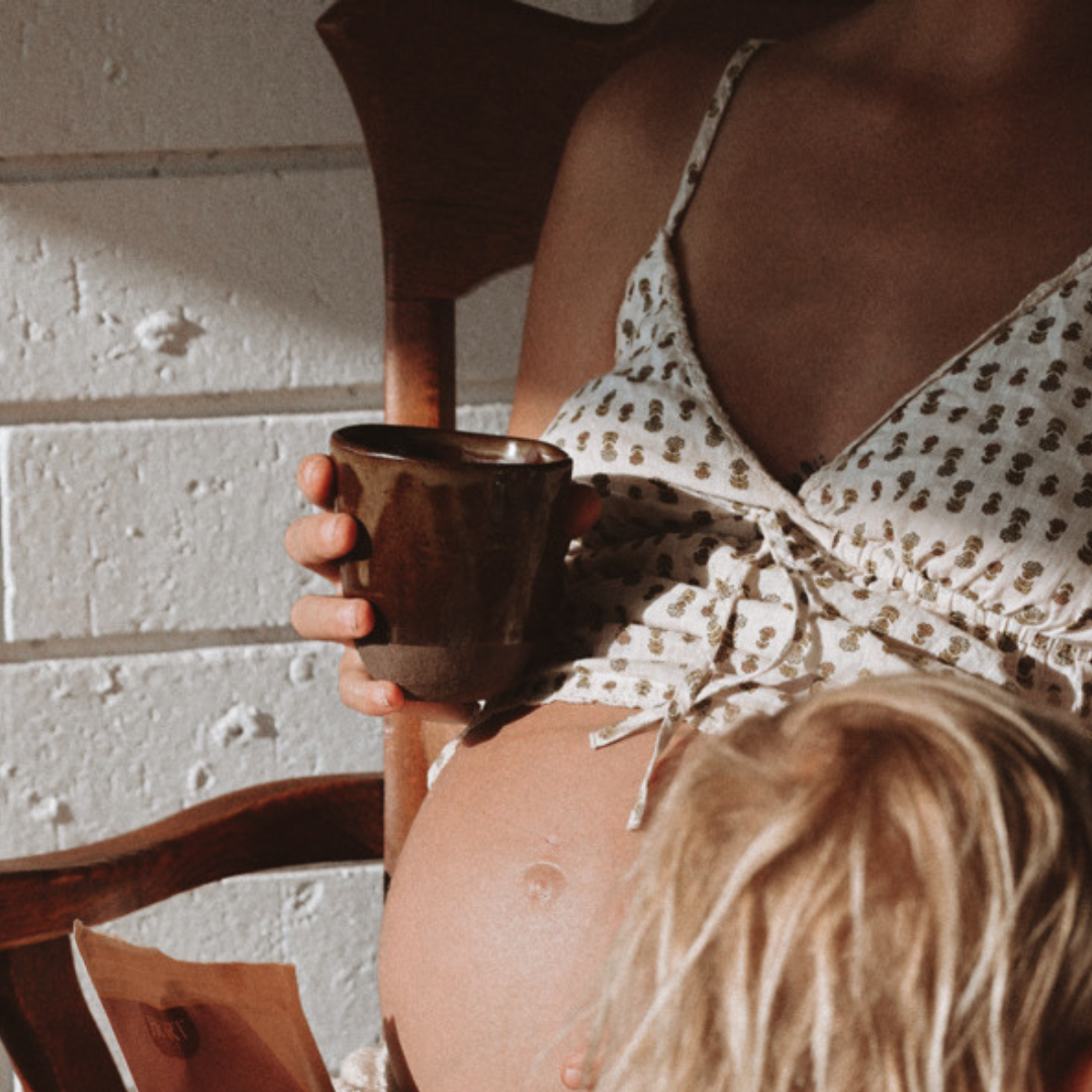 Pregnant woman holding a mug with a child sitting on her lap, against a light-colored wall.