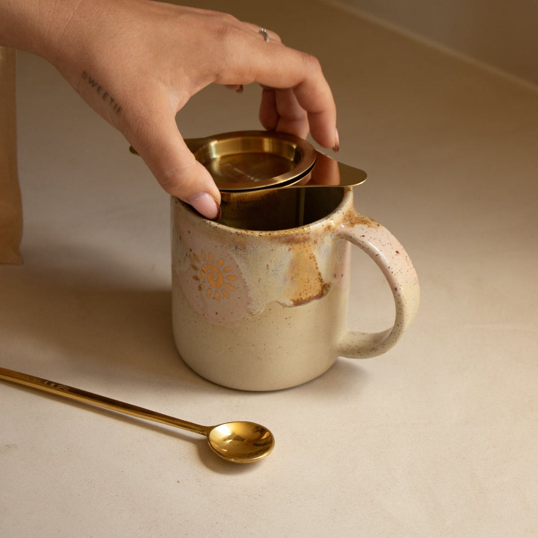 Person placing a gold lid on a ceramic mug with a spoon beside it on a beige surface.