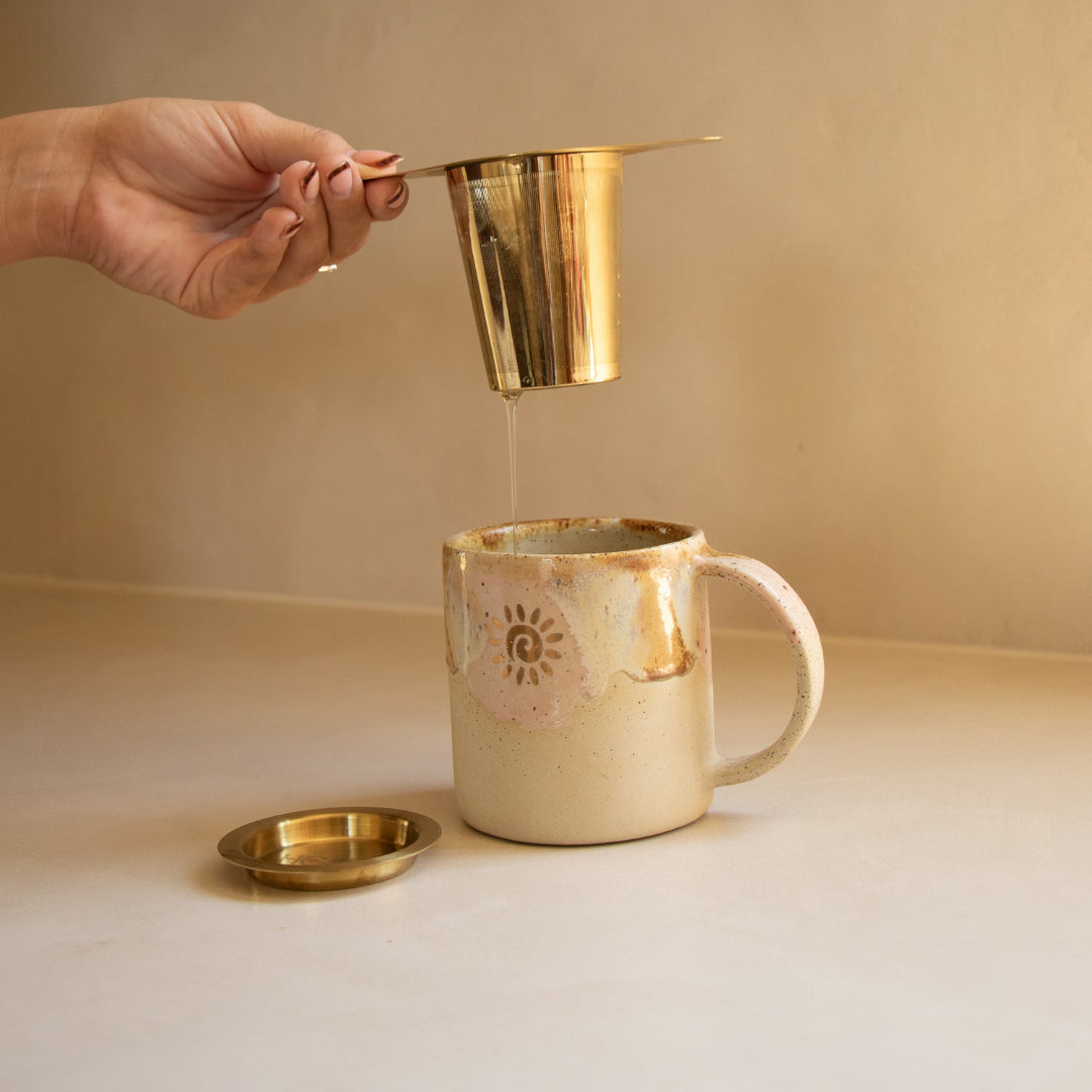 Person pouring liquid from a gold metal container into a ceramic mug on a neutral background
