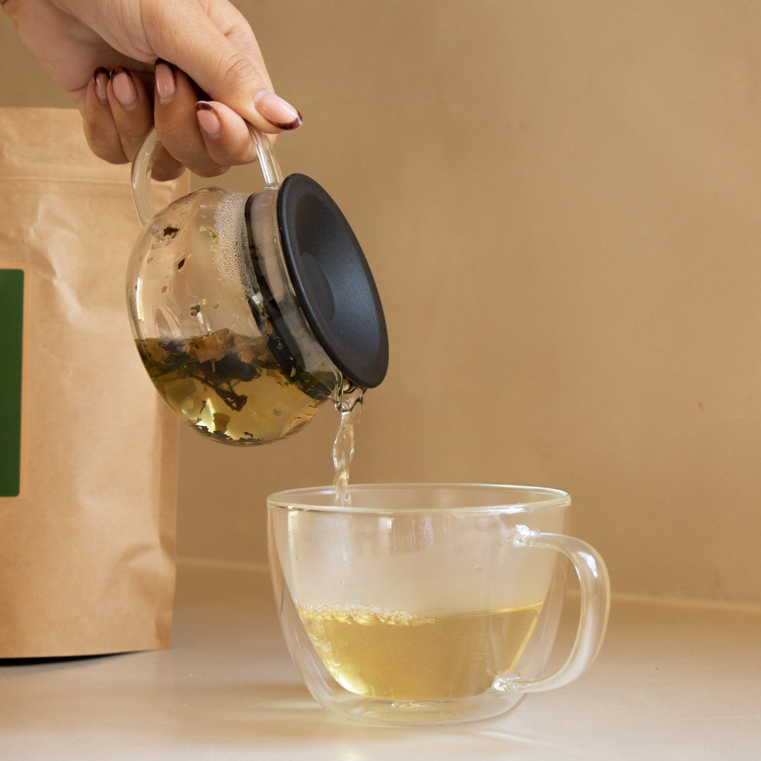 Person pouring tea from a glass teapot into a clear mug with a brown background