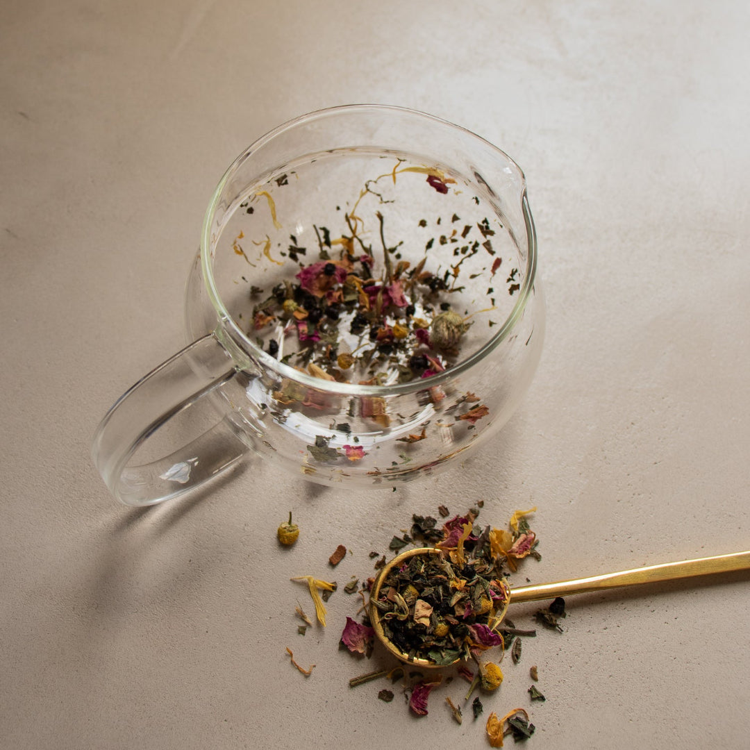 Clear glass mug with dried herbs and a gold spoon on a light surface