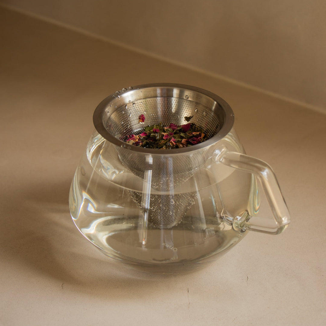 A clear glass teapot with a metal mesh infuser basket containing loose dried flowers, set on a beige surface against a matching beige background.