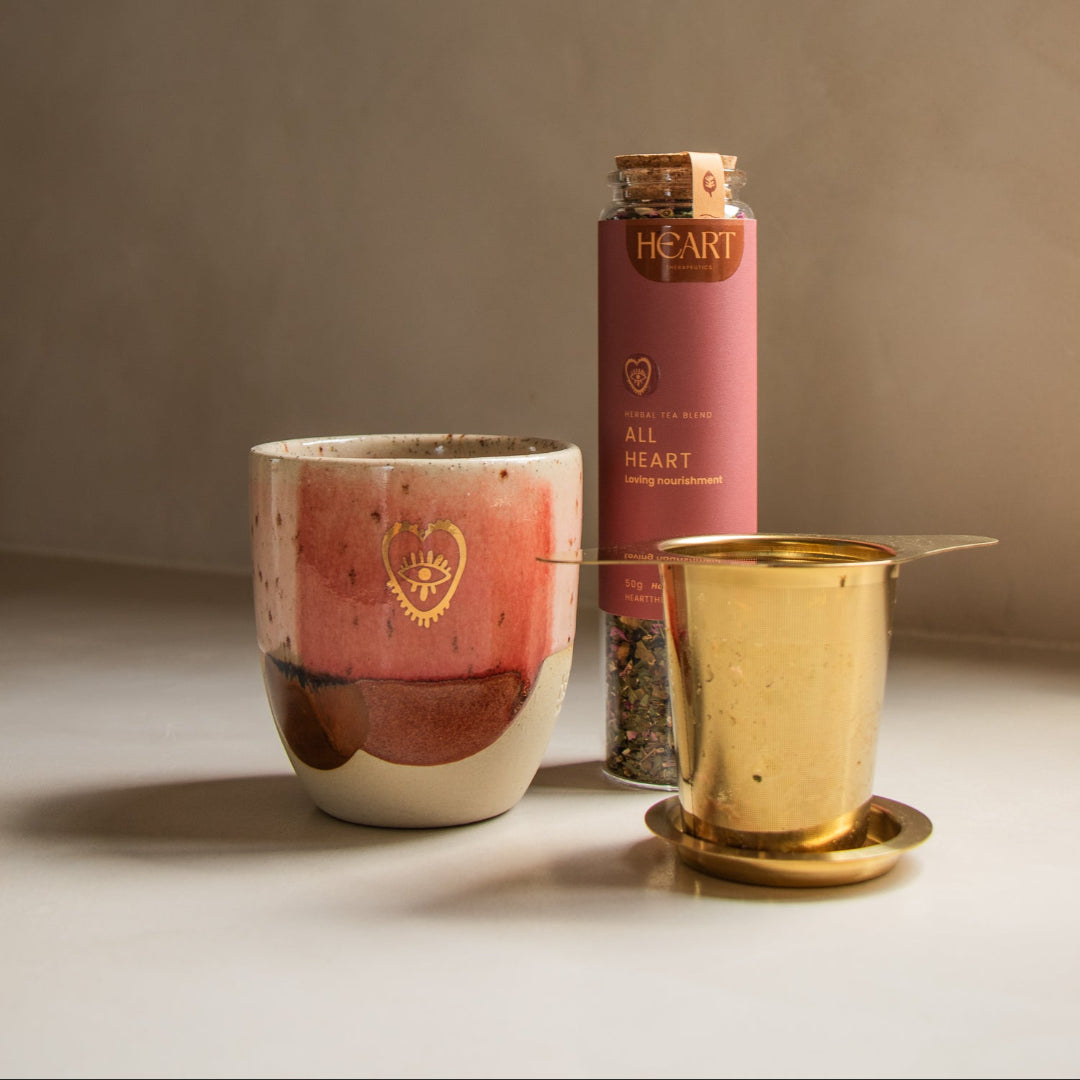 Ceramic mug, pink cylindrical container labeled 'All Heart', and gold metal cup on a neutral background